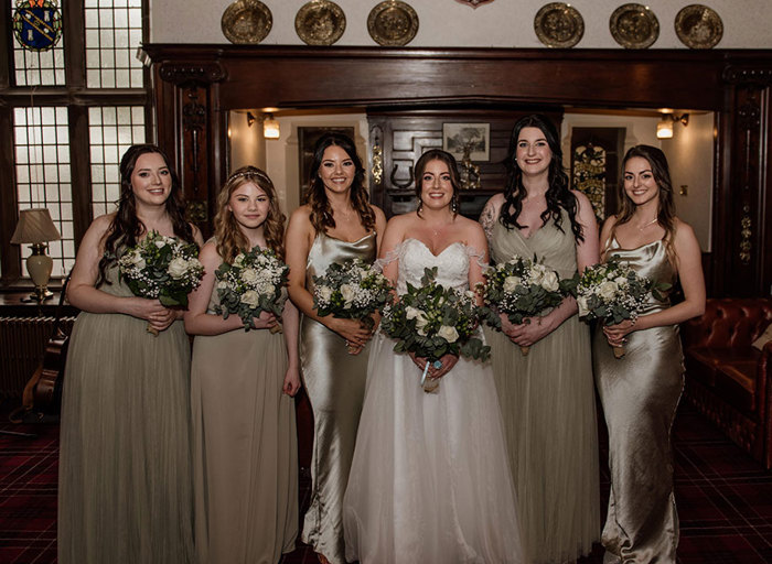 A bride in a white dress stands next to five bridesmaids who are wearing sage green dresses, all of them are holding white bouquets