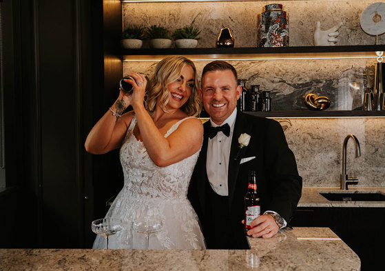 A bride and groom posing for a picture while making a cocktail.