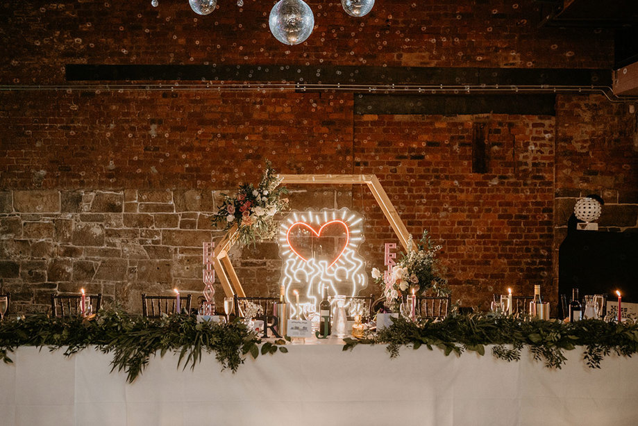 A Table With A White Tablecloth, Greenery And Keith Haring Neon Heart Sign In A Wooden Hexagon