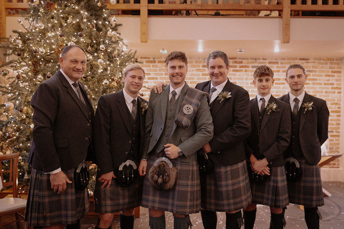 groom in green jacket and red, blue, green kilt with matching fly plaid stands in the middle of five groomsmen in same tartan kilts but darker jackets as they all smile in front of glittering Christmas tree