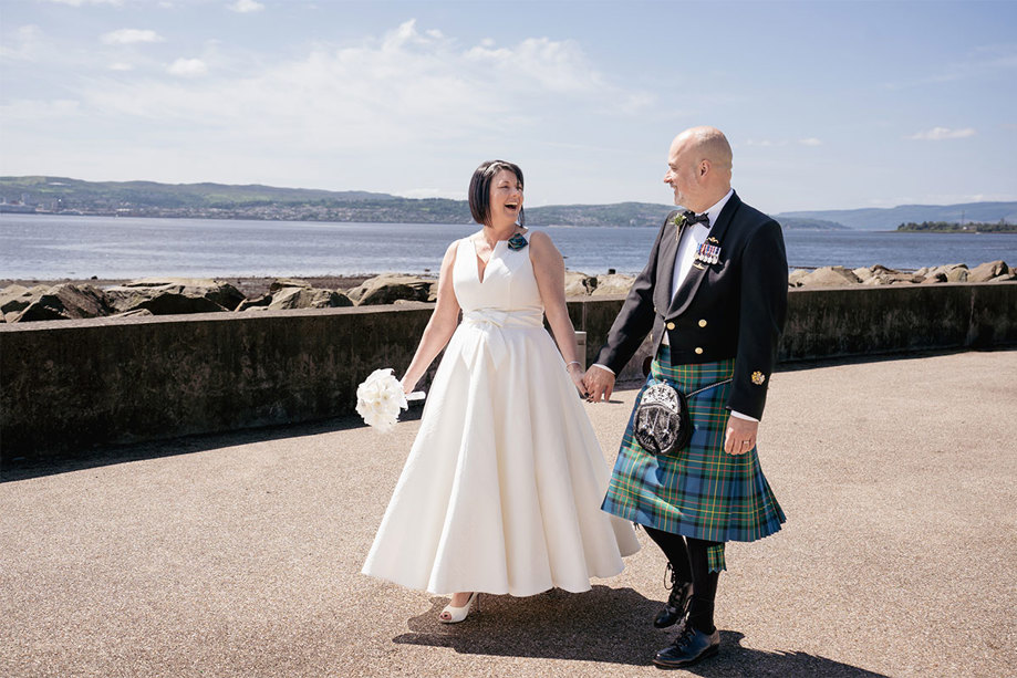 A bride in a wedding dress with an a-line skirt and a groom wearing a green and blue kilt walk hand in hand along a beach front