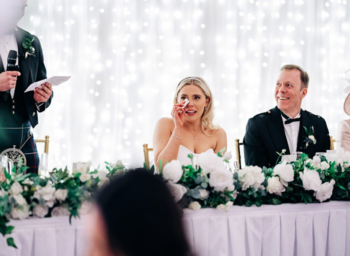 emotional bride dabbing tears away while seated at a long table decorated with white flowers