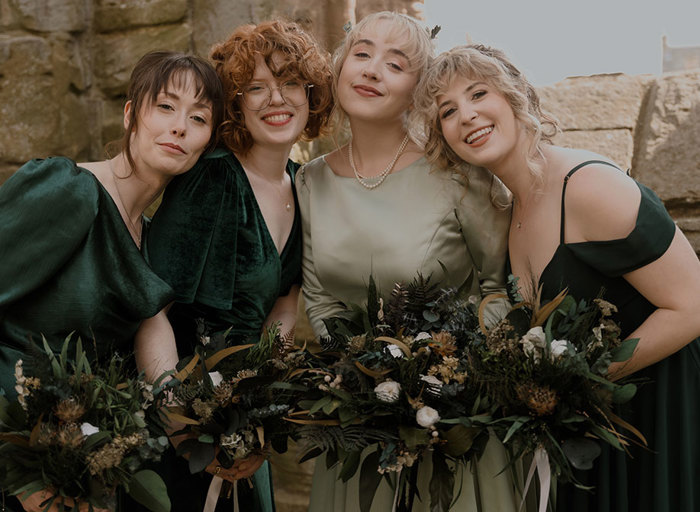 a bride and three bridesmaids wearing green dresses standing in front of a stone wall