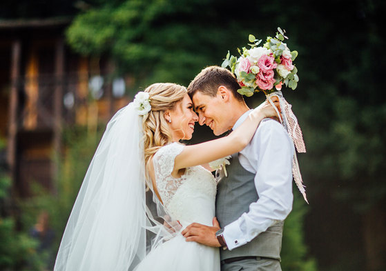 A bride and groom stand with their arms around each other and the bride is holding a white and pink bouquet