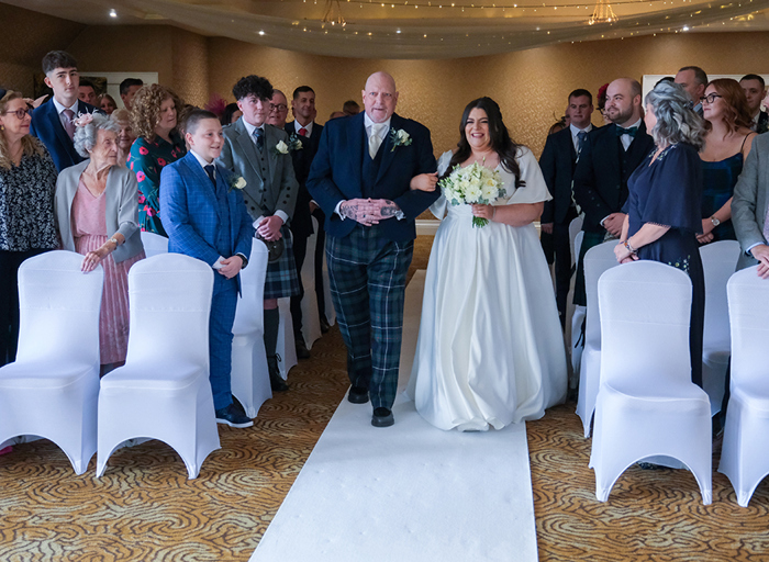 A bride walks down the aisle with her father with the guests on either side of the aisle standing and looking at them