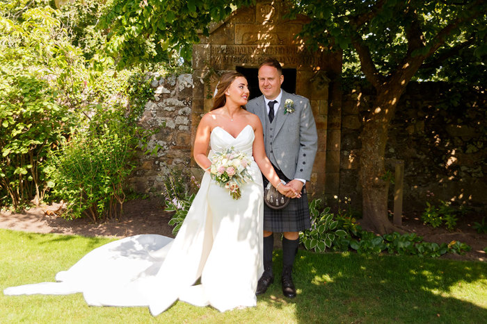 A bride in a long dress and a groom in a kilt stand together in the shade of a tree