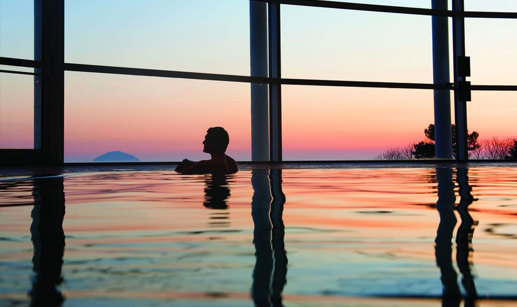 A man in a swimming pool looking out of large windows at a colourful sunset over a hill in the distance