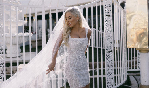 bride in embellished white mini dress poses in front of white gates with her long veil cast out to one side