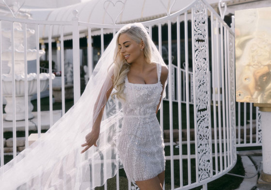 bride in embellished white mini dress poses in front of white gates with her long veil cast out to one side