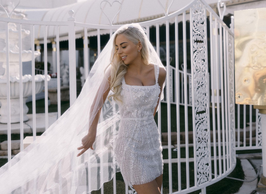 bride in embellished white mini dress poses in front of white gates with her long veil cast out to one side