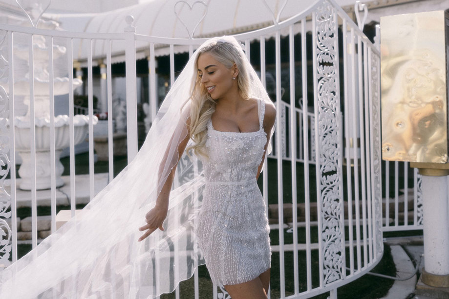 bride in embellished white mini dress poses in front of white gates with her long veil cast out to one side