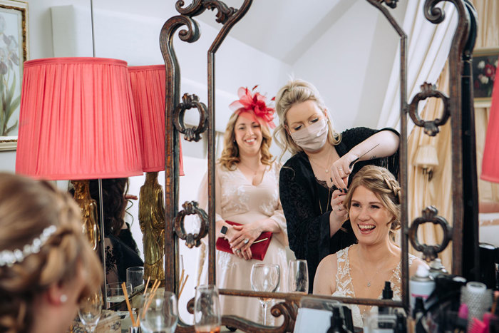 Bride smiling as she gets her hair done with guest watching on smiling