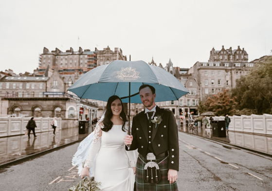 Groom holds umbrella over bride as they walk through Edinburgh