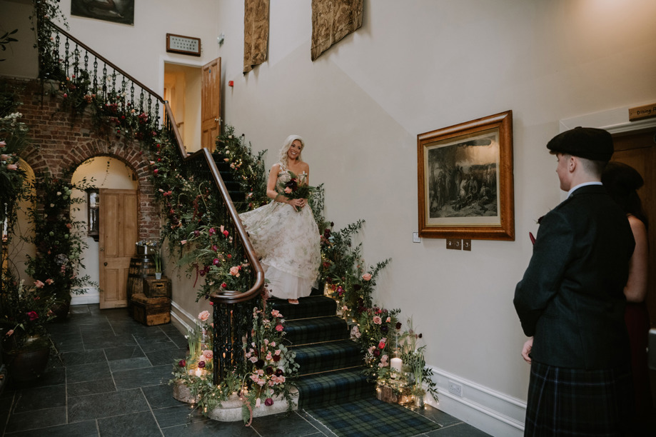 Groom stands at the bottom of staircase as bride walks down towards him smiling in wedding dress at Logie Country House, Aberdeen