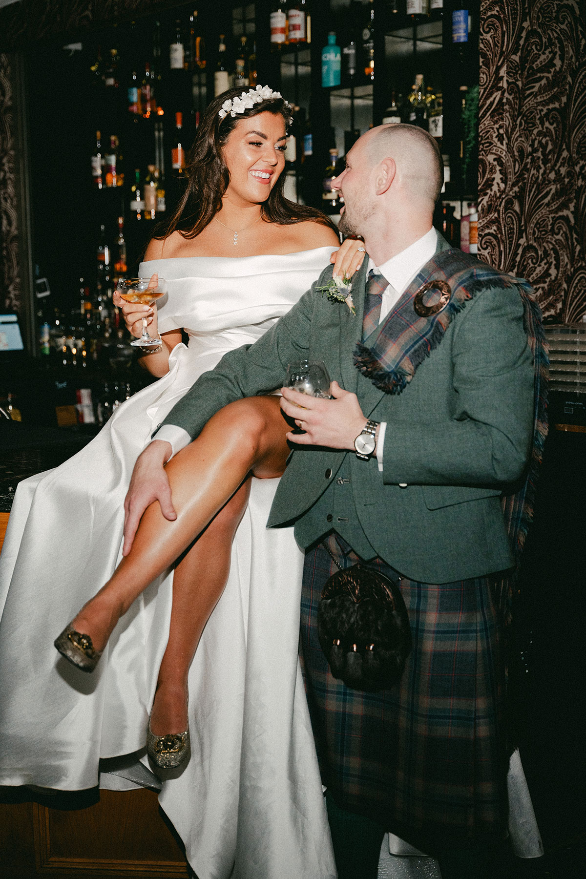 bride in off the shoulder wedding dress and floral headband sits on bar holding a cocktail glass and smiles at groom in green, blue and purple tartan kilt outfit with green jacket who also holds a drinks glass