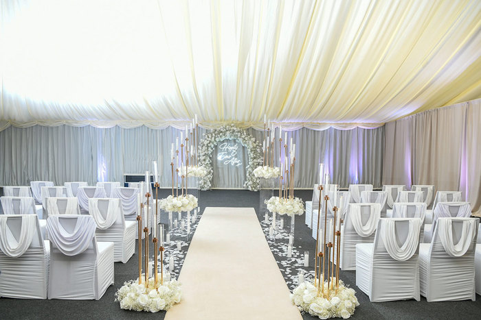 white ceremony space with a cream coloured aisle runner, tall candle holders, and a floral arch at the top of the aisle