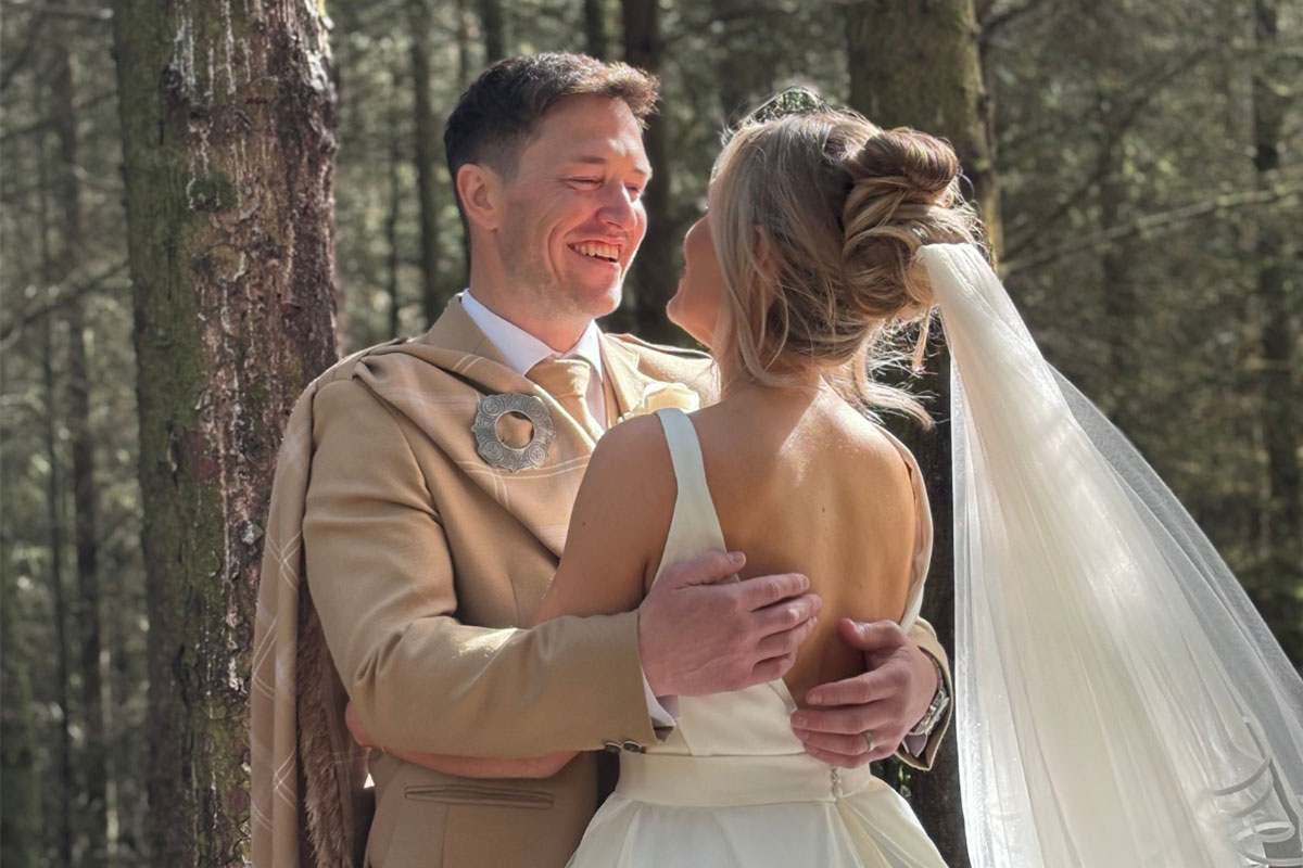 a bride wearing a wedding dress and a groom wearing a beige kilt stand with their arms around each other in the woodlands