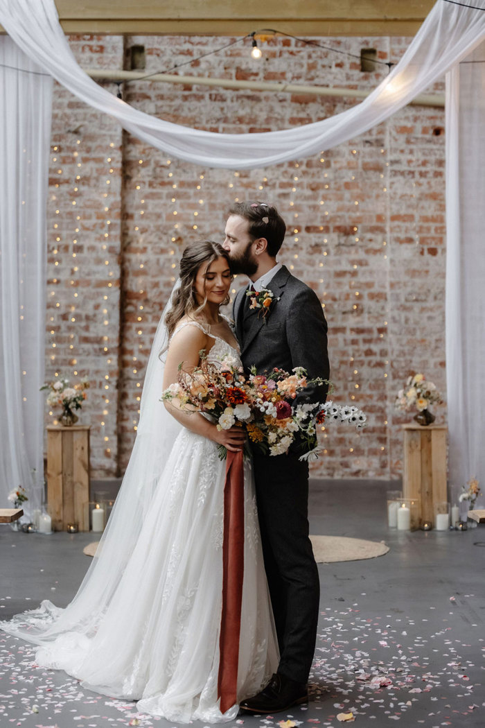 bride and groom stand closely in each other's embrace as groom kisses her on the forehead. white drapes and coloured confetti decorate the room and bride's wildflower bouquet is tied together by long ribbon that trails to the floor