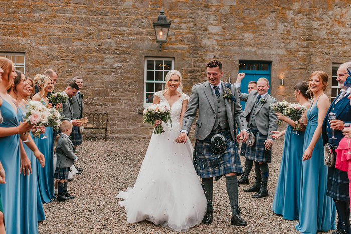 A bride and groom walk through a courtyard past their wedding guests as they throw colourful confetti over the couple