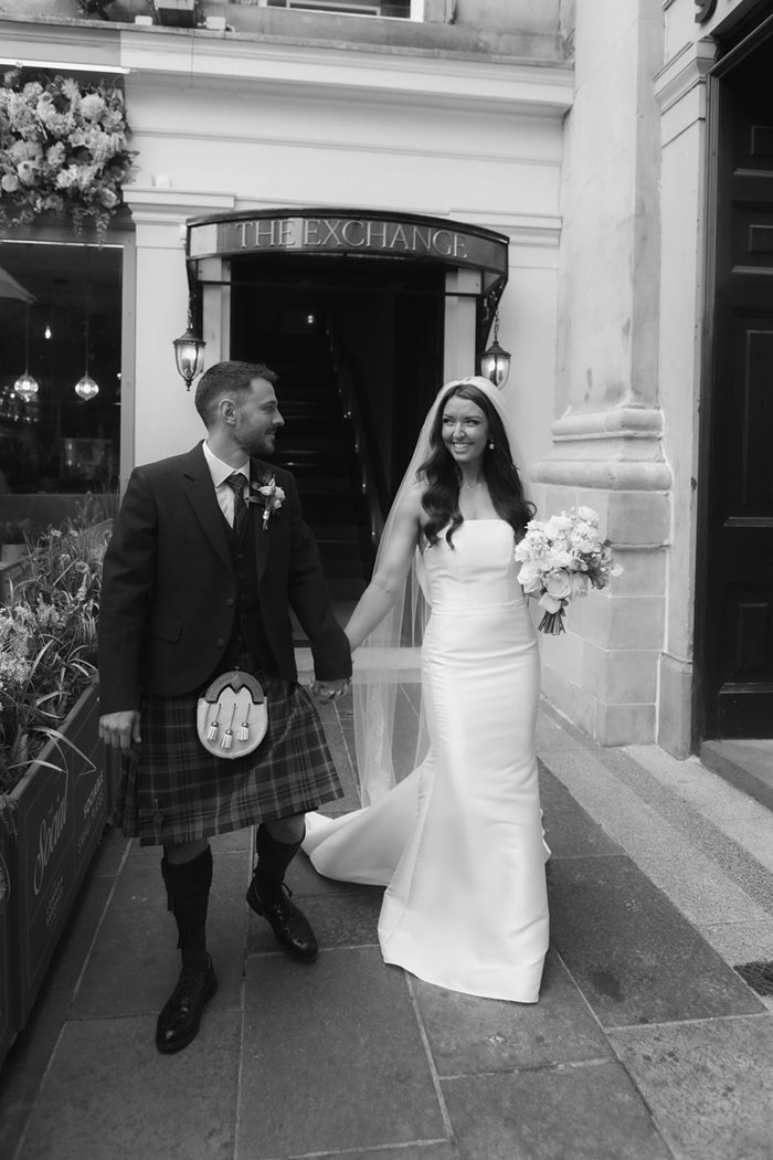 A black and white photo of a bride in a fitted dress and a groom in a kilt holding hands and standing outside of The Exchange