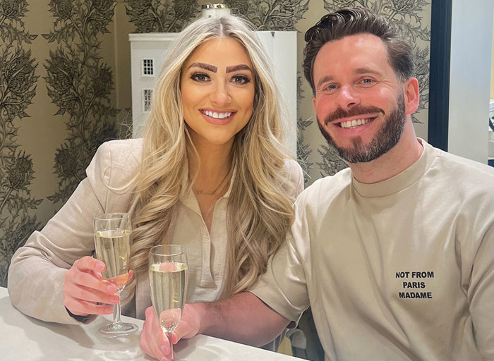 blonde woman smiling next to brunette man smiling, both wearing cream clothing and holding champagne flutes