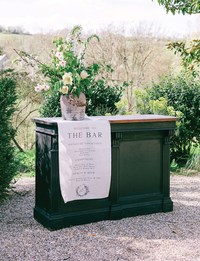 a green standalone bar with a wooden countertop is placed outdoors and decorated with a linen bar sign and a tall vase of flowers