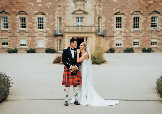 A bride in a wedding dress and a groom in a kilt standing in front of Archerfield House.