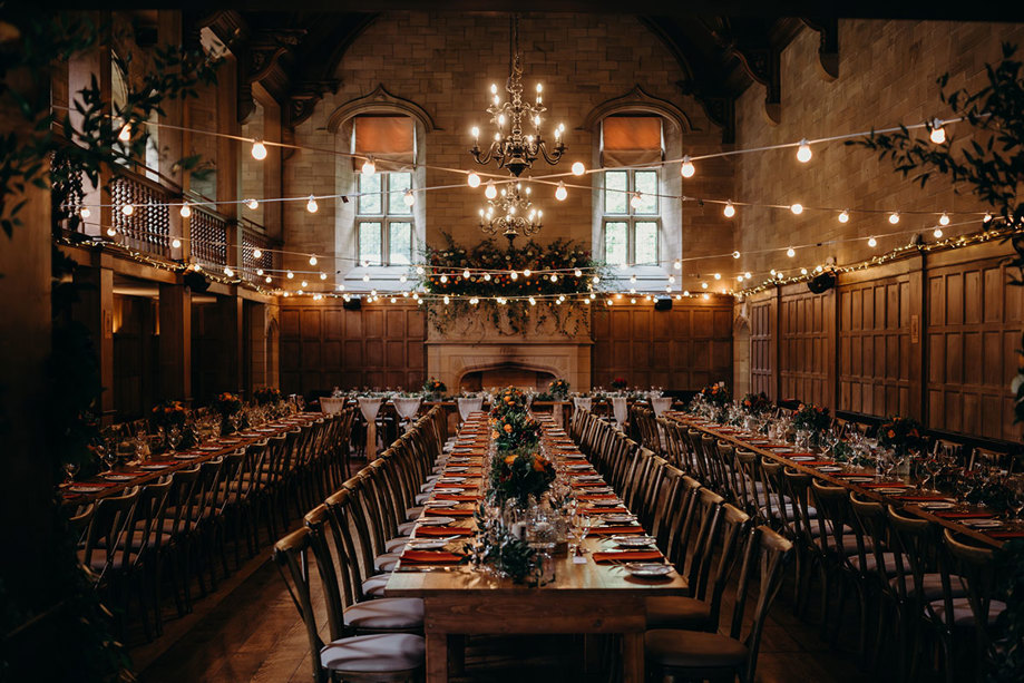 the ballroom at Achnagairn Castle with rows of long tables and chairs. There is a large stone fireplace in the background and strings of lights hanging above the tables.