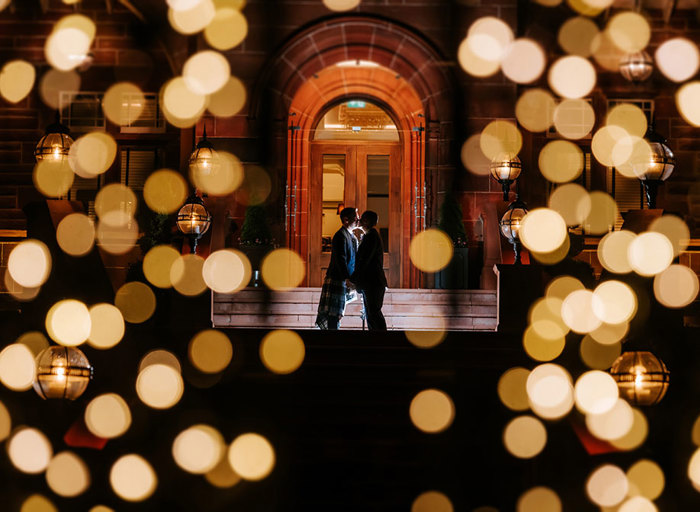 a backlit silhouetted image of two grooms kissing in an arched doorway with dappled spots of lighting in foreground