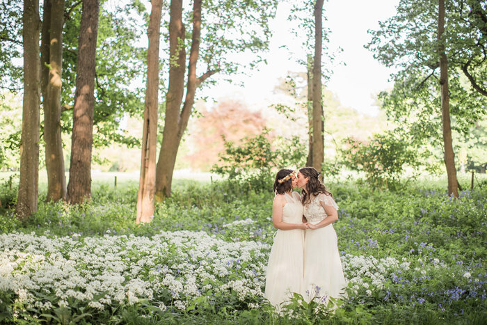 two brides kissing in a woodland with lots of white and blue flowers by their feet.