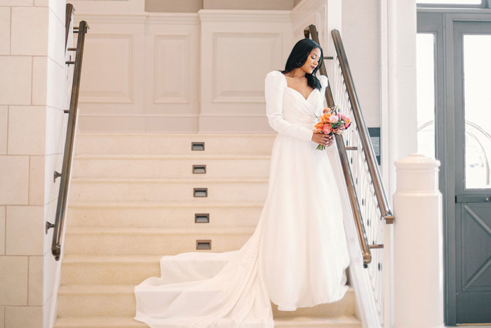 a bride in a long sleeved trailing gown stands on a staircase holding a pink and orange bouquet