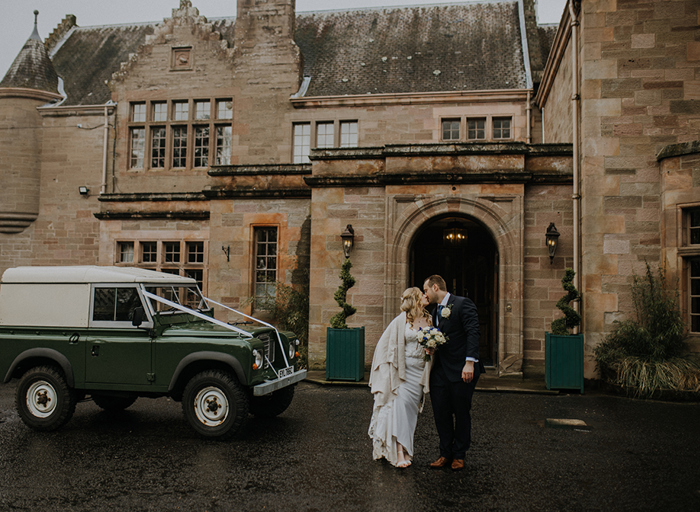 A bride and groom stand in front of a country house entrance and kiss with a green jeep next to them