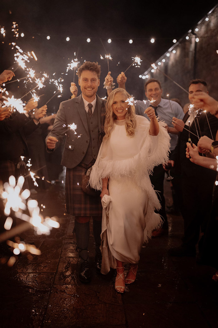blonde bride in feathery cape and white wedding dress holds end of dress in one hand and sparkler in the other as she walks side by side with groom in green jacket and red, blue, green kilt with matching fly plaid, also holding a sparkler 