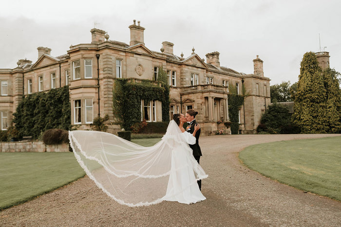 Bride and groom kiss outside grand brick venue with bride's veil blowing in the breeze