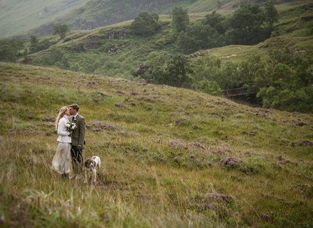 a bride and groom kissing in the heather covered hills of Glencoe in Scotland. They have a spaniel dog on a lead with them