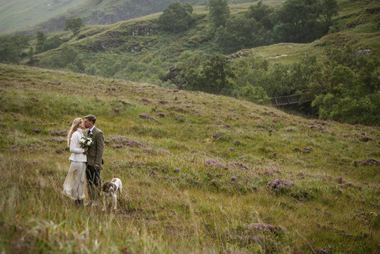 a bride and groom kissing in the heather covered hills of Glencoe in Scotland. They have a spaniel dog on a lead with them