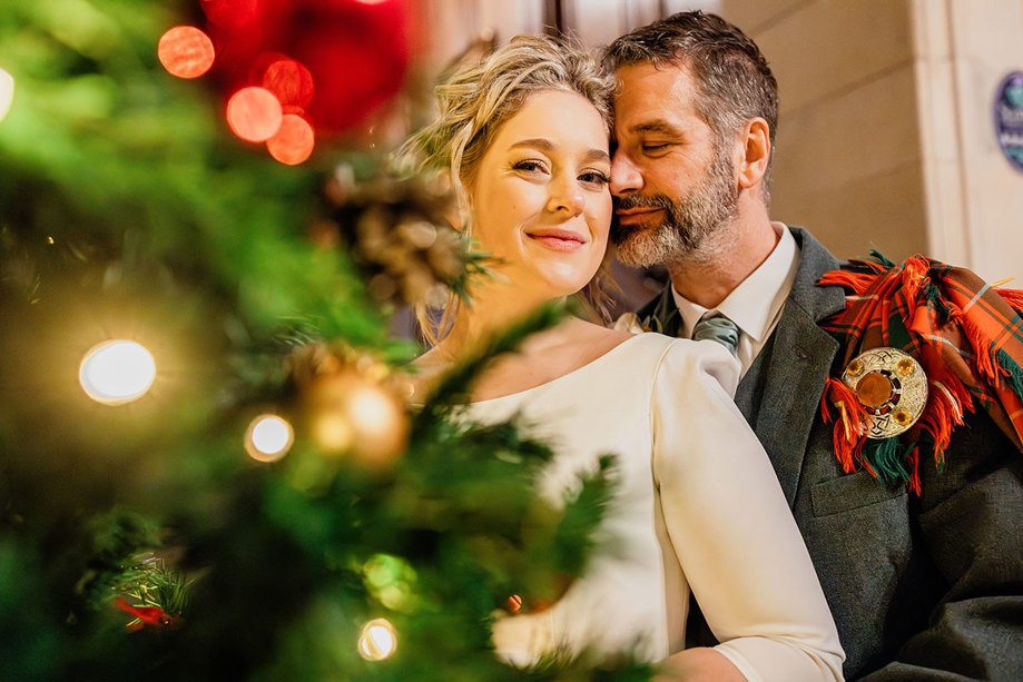 a close up portrait of bride in white wedding dress and groom in red and green tartan outfit as they stand directly behind a the branches of a christmas tree