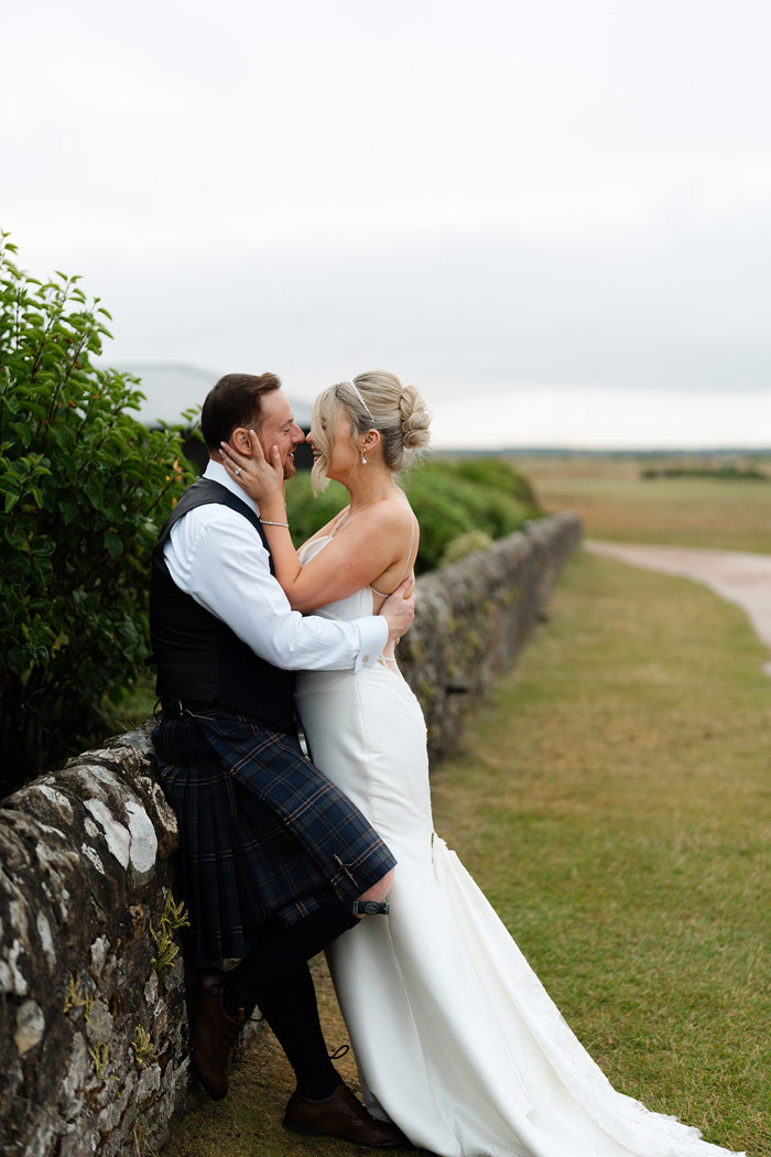 a groom leaning against a stone wall with bride holding his face tenderly. They are on the Old Course golf course in St Andrews.