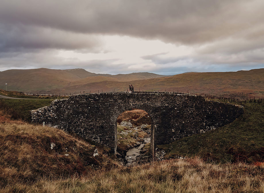 a bride and groom standing on an old black stone bridge surrounded by autumn hills and countryside