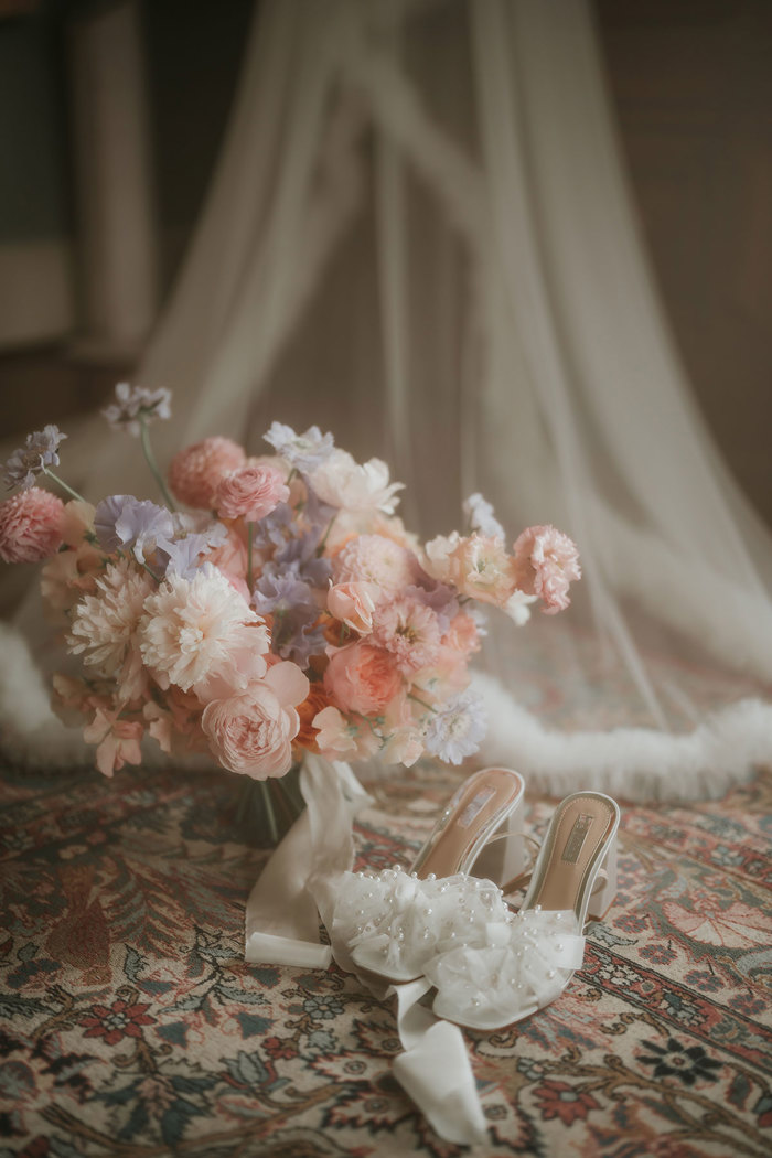 A pair of white high heels with tulle bows sitting next to a pink and purple bouquet, which are both on the floor in front of a long veil 