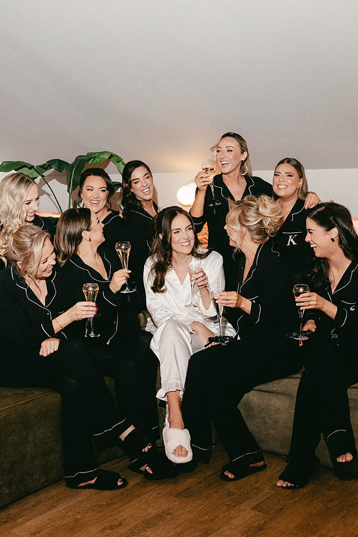 bride in long-sleeve white pyjamas surrounded by nine women in black long-sleeve pyjamas all hold glasses of prosecco while sitting on a green sofa