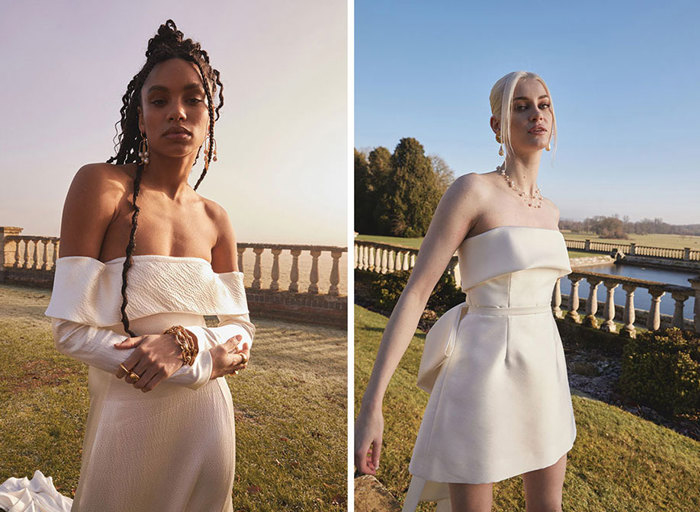 Two women in wedding dresses standing outside on grass next to a wall of pillars