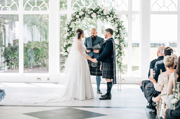 Bride and groom hold hands during wedding ceremony with white floral arch behind them