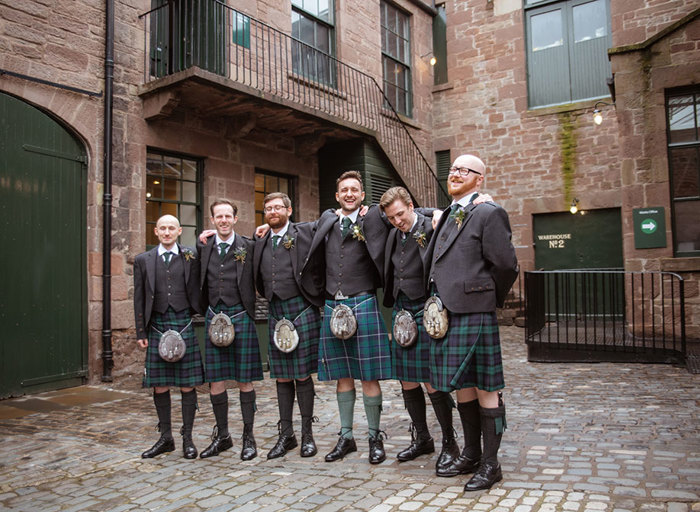 six men in kilts standing on a cobbled pavement surrounded by brown brick buildings