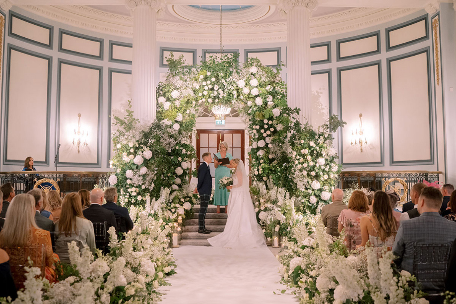 Bride and groom have white flower installation behind them during ceremony