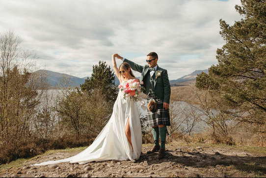 A Bride And Groom Dancing on a stony surface with trees, a lake and countryside in the background