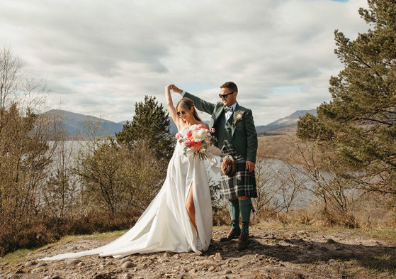 A Bride And Groom Dancing on a stony surface with trees, a lake and countryside in the background