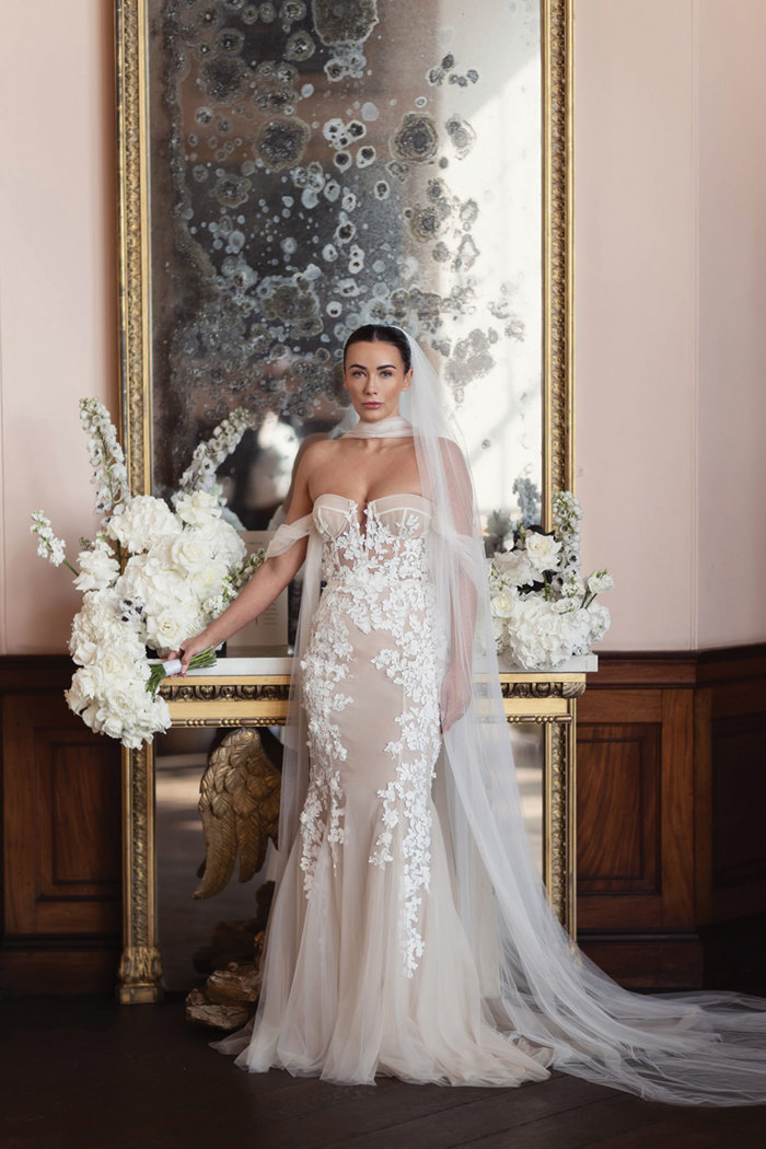 a bride standing in front of an ornate gold antique mirror with white bouquet and floral arrangements.