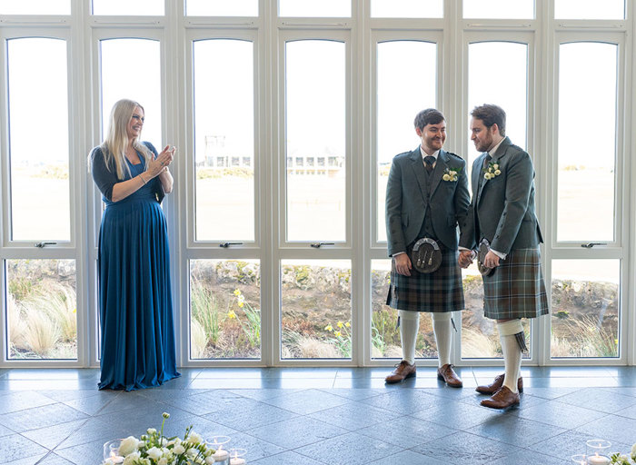 a person wearing a long blue dress clapping as two men wearing green tweed kilt outfits hold hands. They are standing in front of tall white floor to ceiling windows on a grey tiled floor