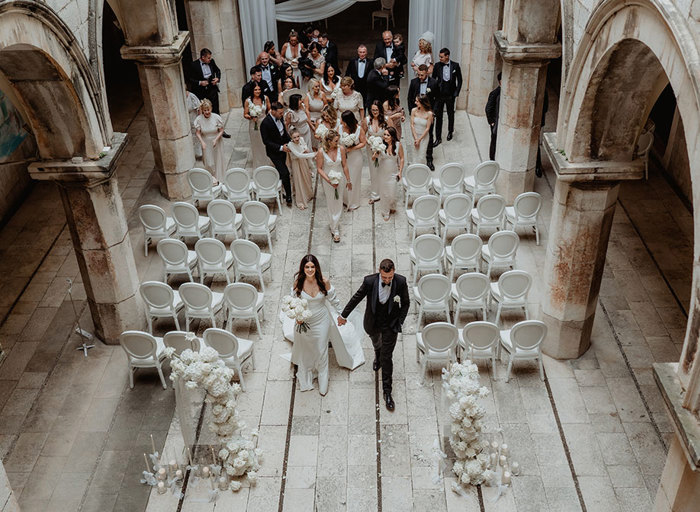 an aerial image of a bride and groom walking hand-in-hand through Sponsa Palace in Dubrovnik. There are white chairs set out in rows either side and the rest of the wedding party stand behind them. 